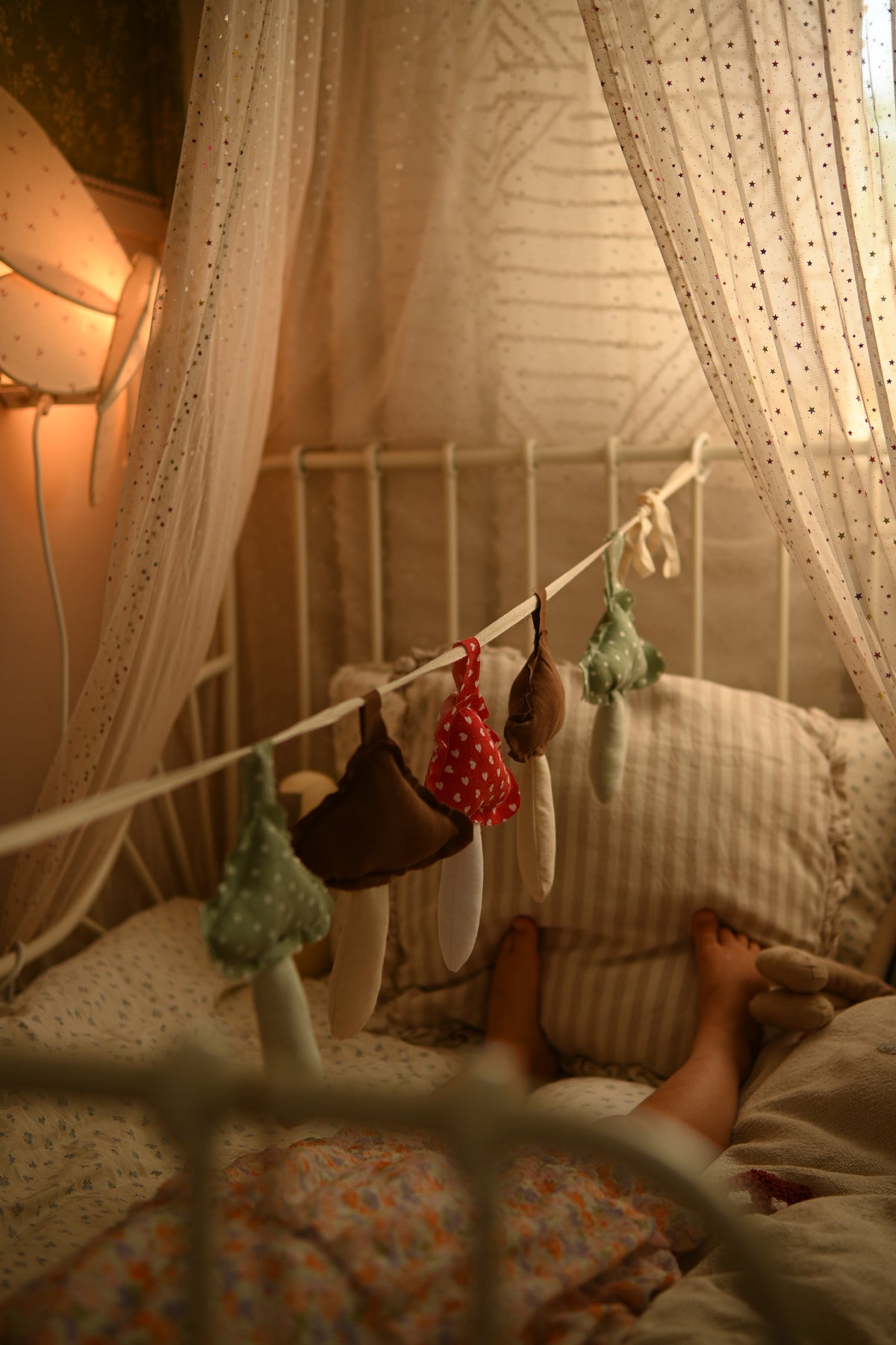 A cozy bedroom scene with a bed canopy and sheer curtains, a string of handmade fabric items hanging across the bed, and a person lying in bed with their feet visible.