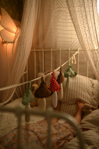 A cozy bedroom scene with a white metal bed frame draped in sheer curtains, decorated with small hanging fabric ornaments. A person is lying in bed under the canopy, with warm ambient lighting and a butterfly wall lamp visible.