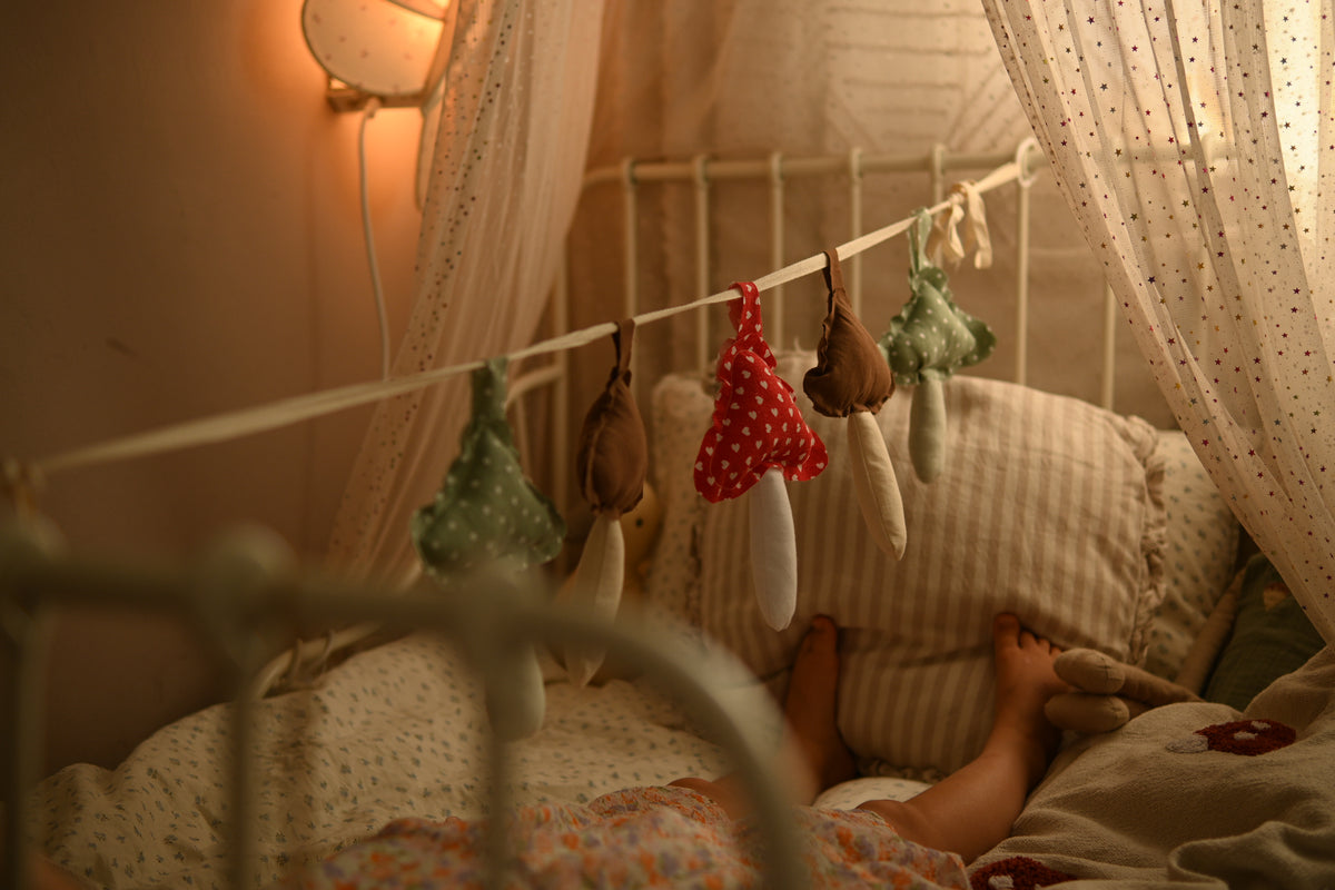 A cozy bedroom scene with a child lying in bed under a white canopy, surrounded by soft bedding and a string of small mushroom-shaped hanging toys.