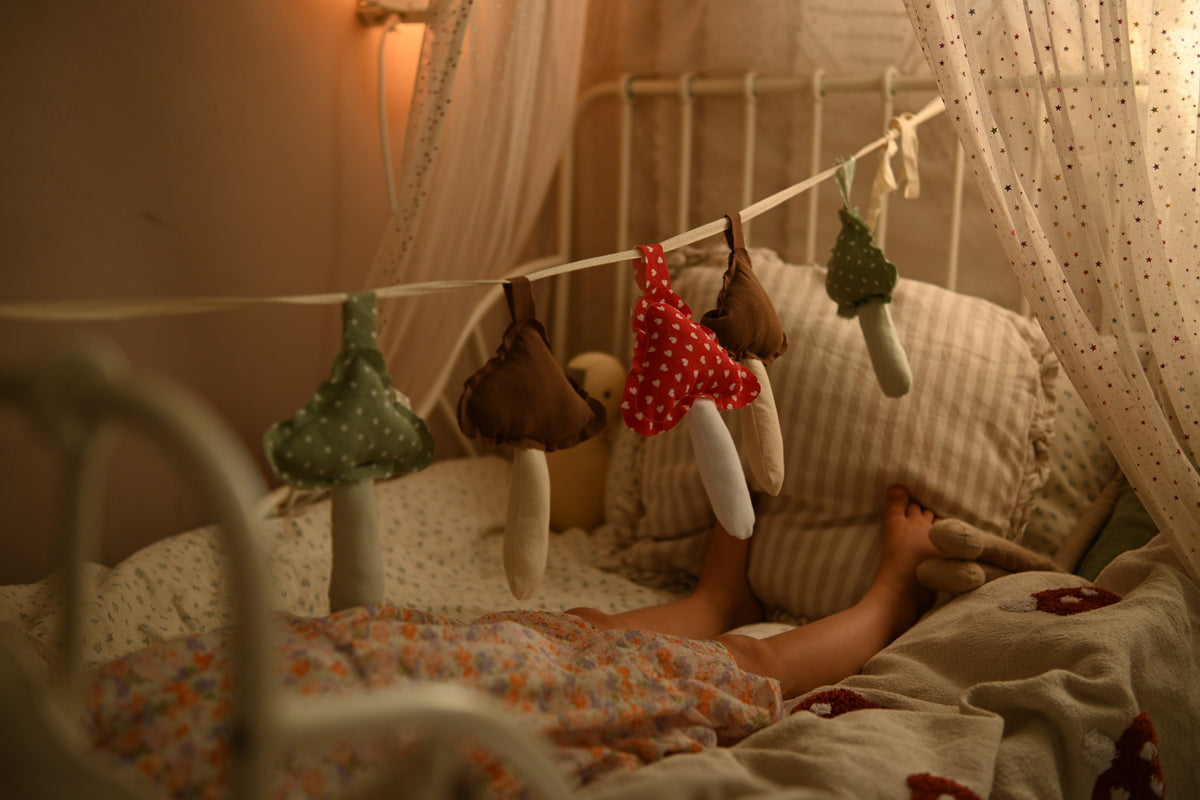 A cozy bedroom scene with a child lying in bed under a white canopy, surrounded by soft blankets and pillows. Hanging on a string are several mushroom-shaped fabric toys in muted green, brown, and red-and-white polka dot colors.