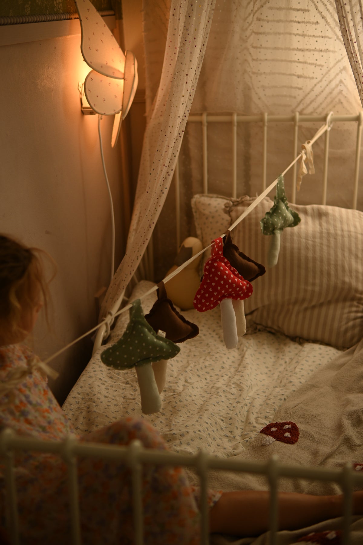 A cozy nursery scene with a child sitting beside a bed under a canopy. The bed is decorated with a string of mushroom-shaped hanging ornaments in green, red, brown, and beige, with a warm fairy light glowing on the wall.