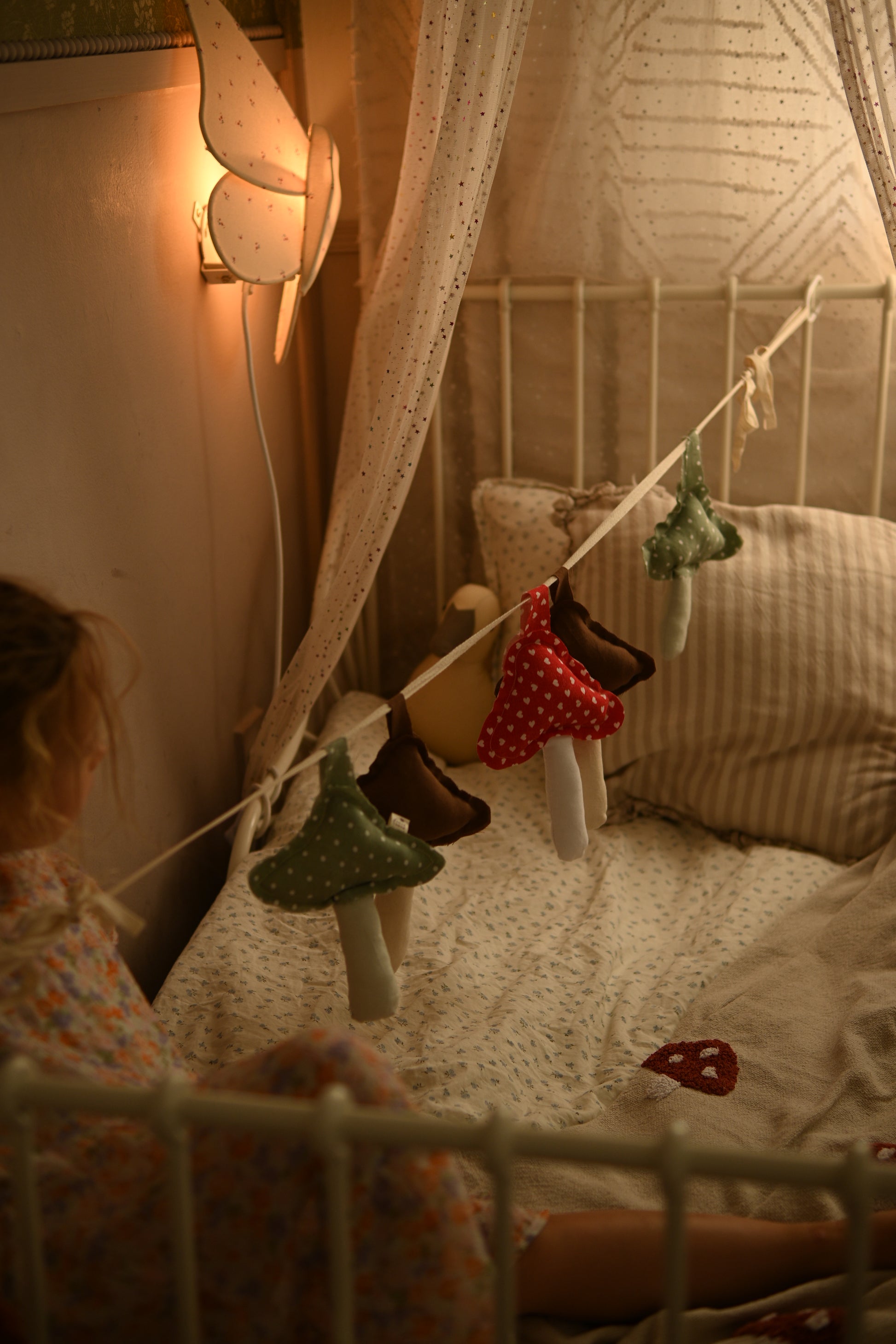 A cozy nursery scene with a child sitting beside a bed under a canopy. The bed is decorated with a string of mushroom-shaped hanging ornaments in green, red, brown, and beige, with a warm fairy light glowing on the wall.