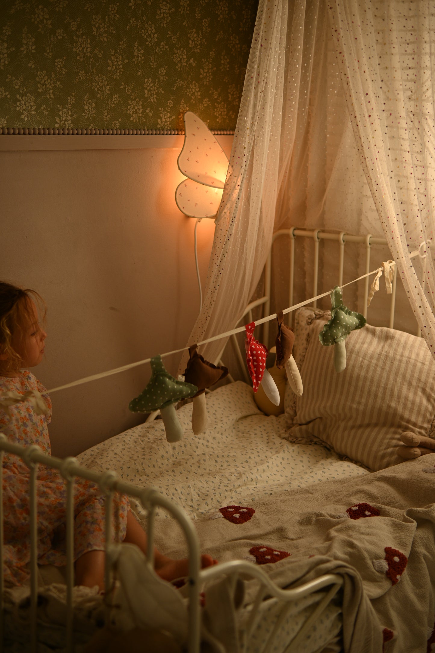 A cozy bedroom scene with a young child sitting on a bed in a white metal frame, surrounded by a sheer canopy, patterned wallpaper, a butterfly-shaped wall lamp, and hanging fairy lights and small plush toys.