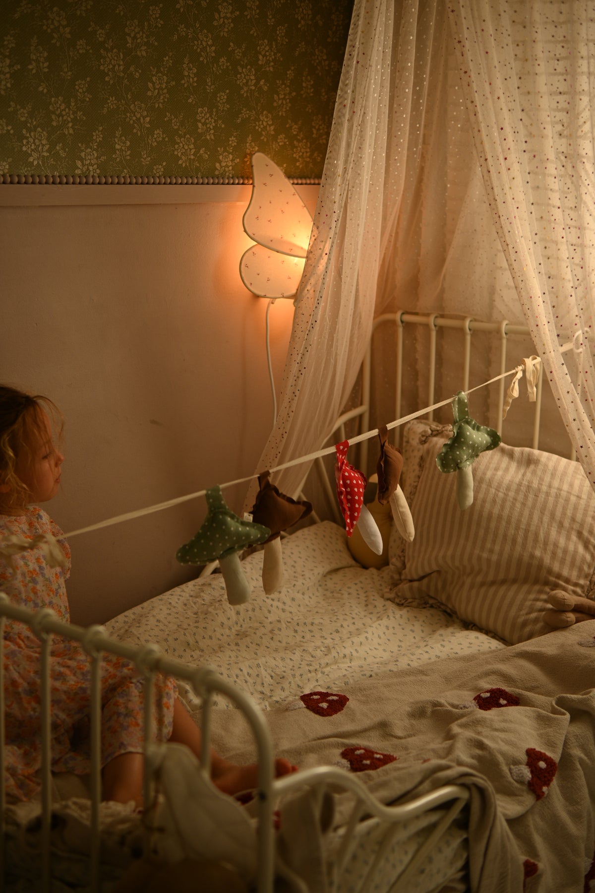A cozy bedroom scene with a young child sitting on a bed in a white metal frame, surrounded by a sheer canopy, patterned wallpaper, a butterfly-shaped wall lamp, and hanging fairy lights and small plush toys.