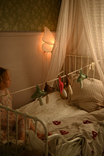 A cozy bedroom scene with a young child sitting on a bed in a white metal frame, surrounded by a sheer canopy, patterned wallpaper, a butterfly-shaped wall lamp, and hanging fairy lights and small plush toys.