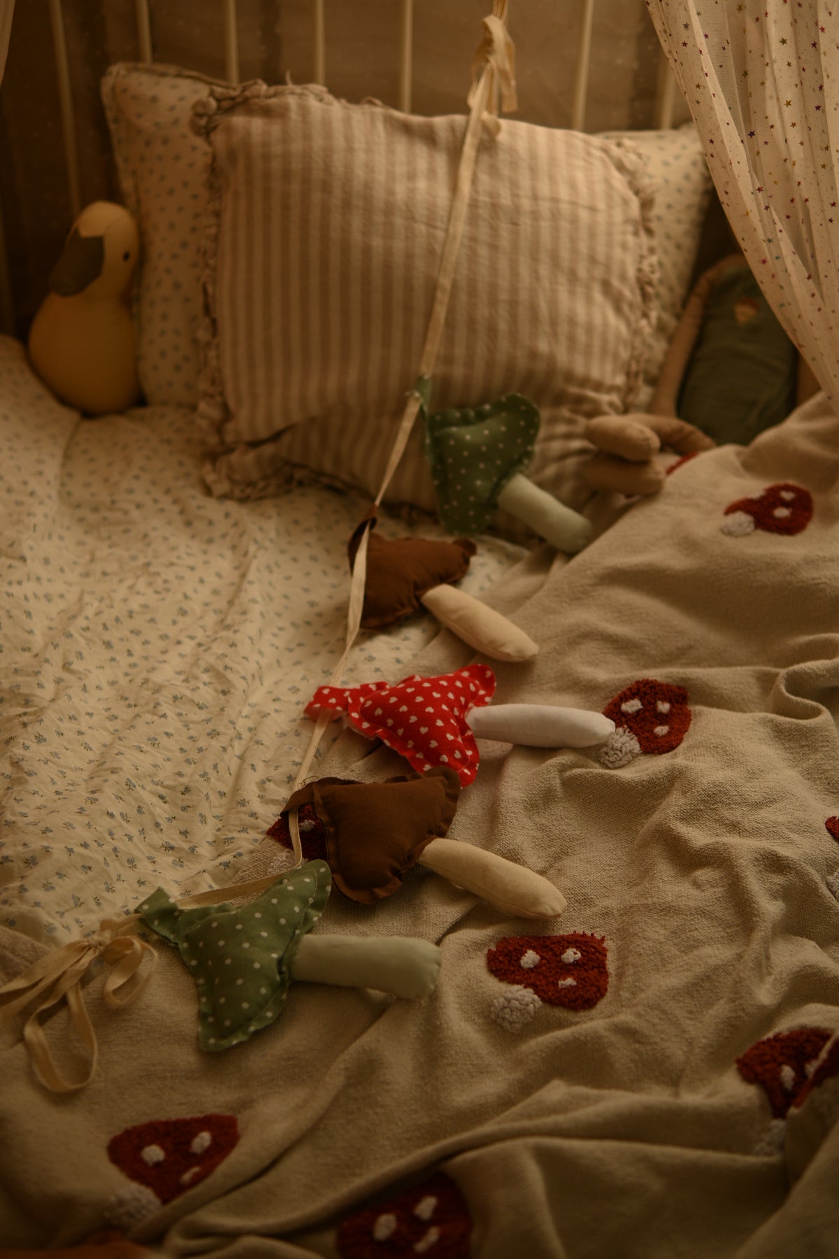 A cozy nursery scene with a bed made up in soft beige fabrics, pillows, and a blanket featuring red mushroom prints. A hanging fabric mobile with small felt mushroom shapes in green, red, and brown is draped across the bed.