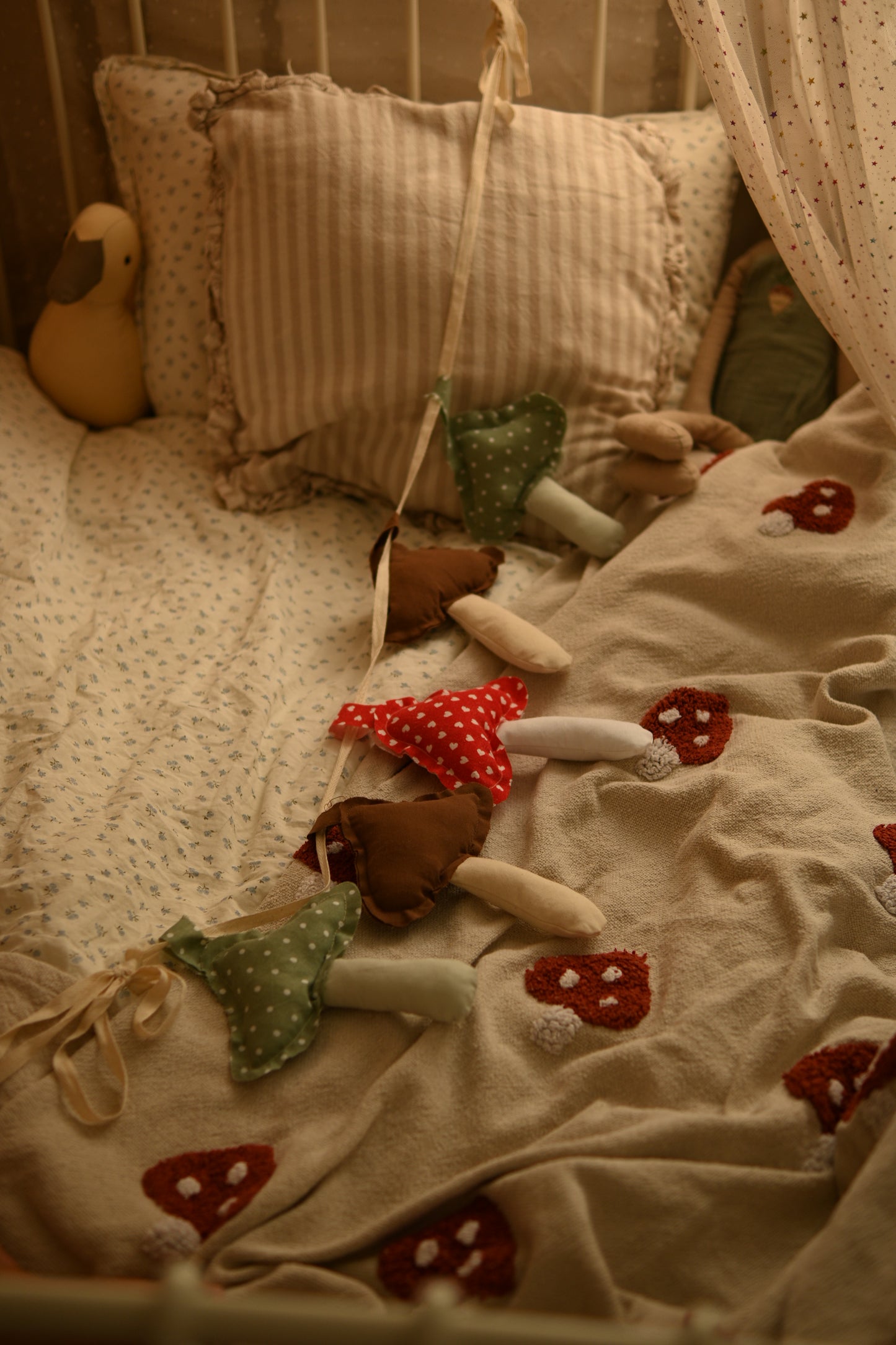 A cozy nursery scene with a bed covered in a beige patterned blanket and matching pillow, decorated with a hanging fabric mushroom garland in red, green, and brown.