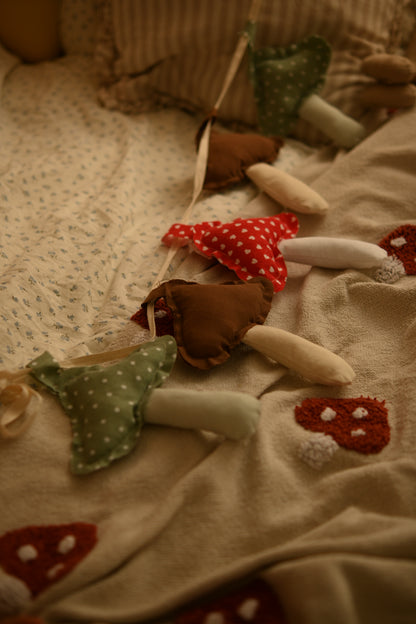 A cozy close-up of a handmade mushroom garland draped over a beige blanket with embroidered red and white mushrooms. The fabric mushrooms are made from brown and green fabrics with white polka dots, red polka dots, and solid colors, with cream-colored stems.