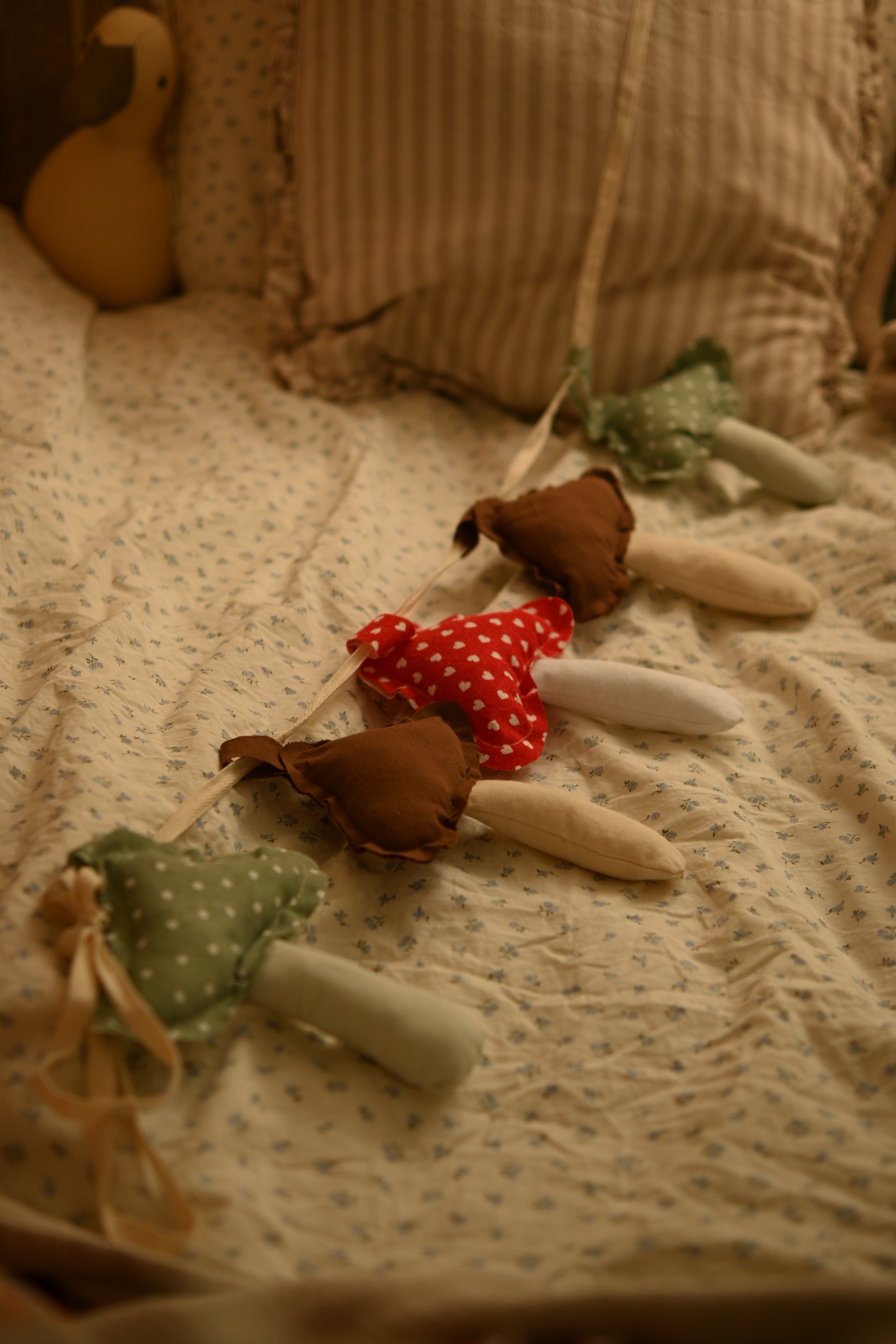 A soft fabric garland of mushroom-shaped ornaments draped across a quilted bed. The garland features brown and green fabric mushrooms with red and white polka-dot centers, set against a warm, cozy bedroom background.