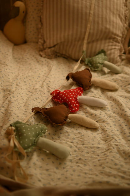 A soft fabric garland of mushroom-shaped ornaments draped across a quilted bed. The garland features brown and green fabric mushrooms with red and white polka-dot centers, set against a warm, cozy bedroom background.