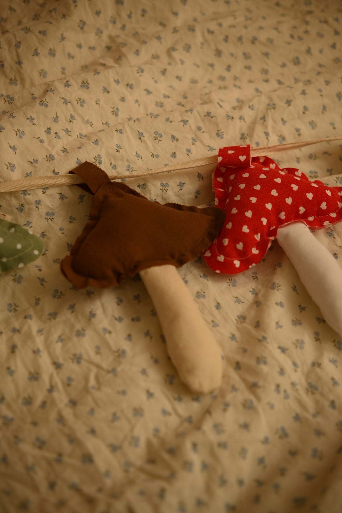 A close-up of a handmade fabric mushroom garland resting on a beige floral baby blanket. The garland features felt mushrooms in brown, red with white hearts, and green, all with cream-colored stems.